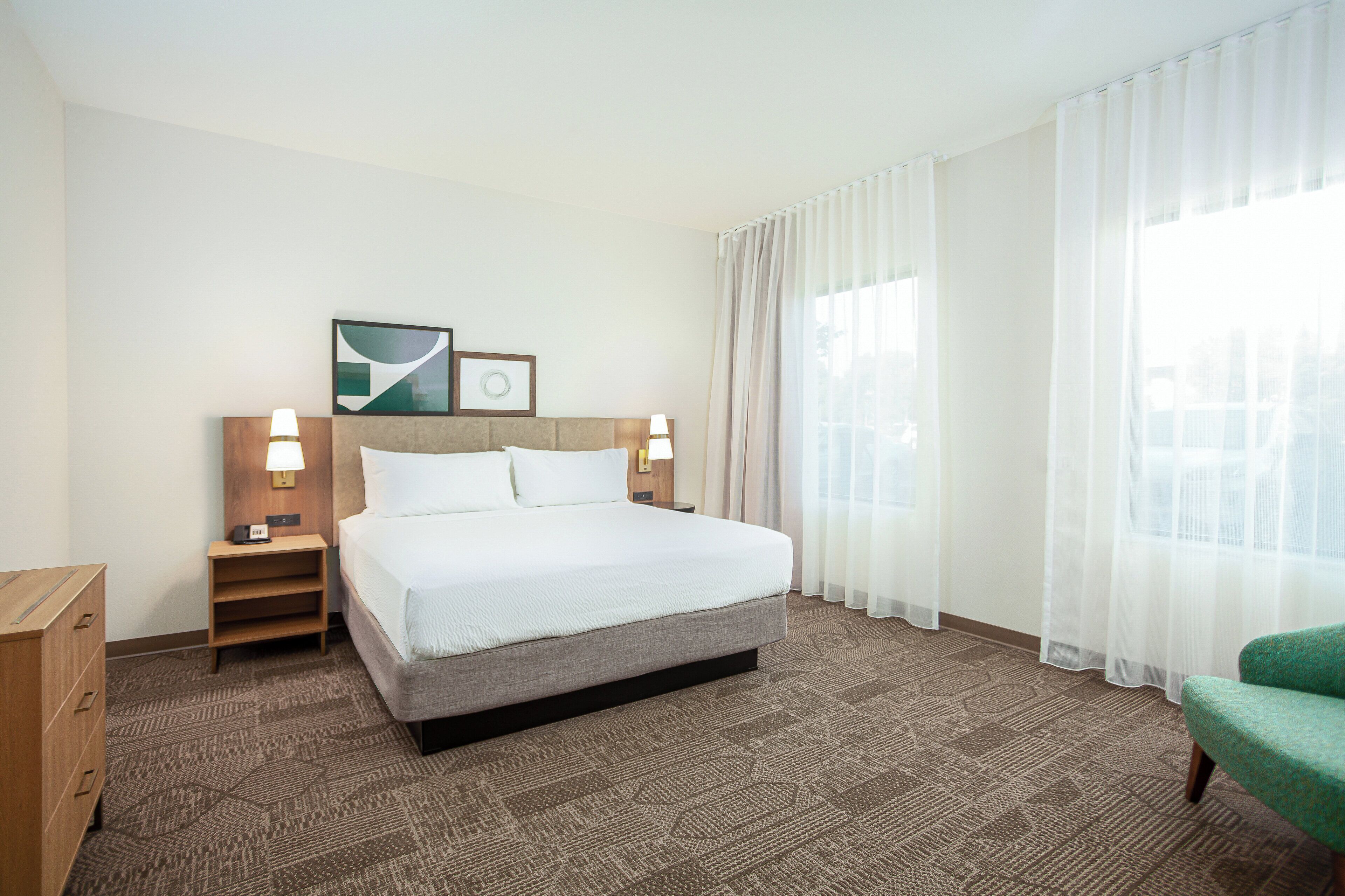 Una habitación de hotel con cama, mesitas de noche, una silla verde y una ventana con cortinas translúcidas.