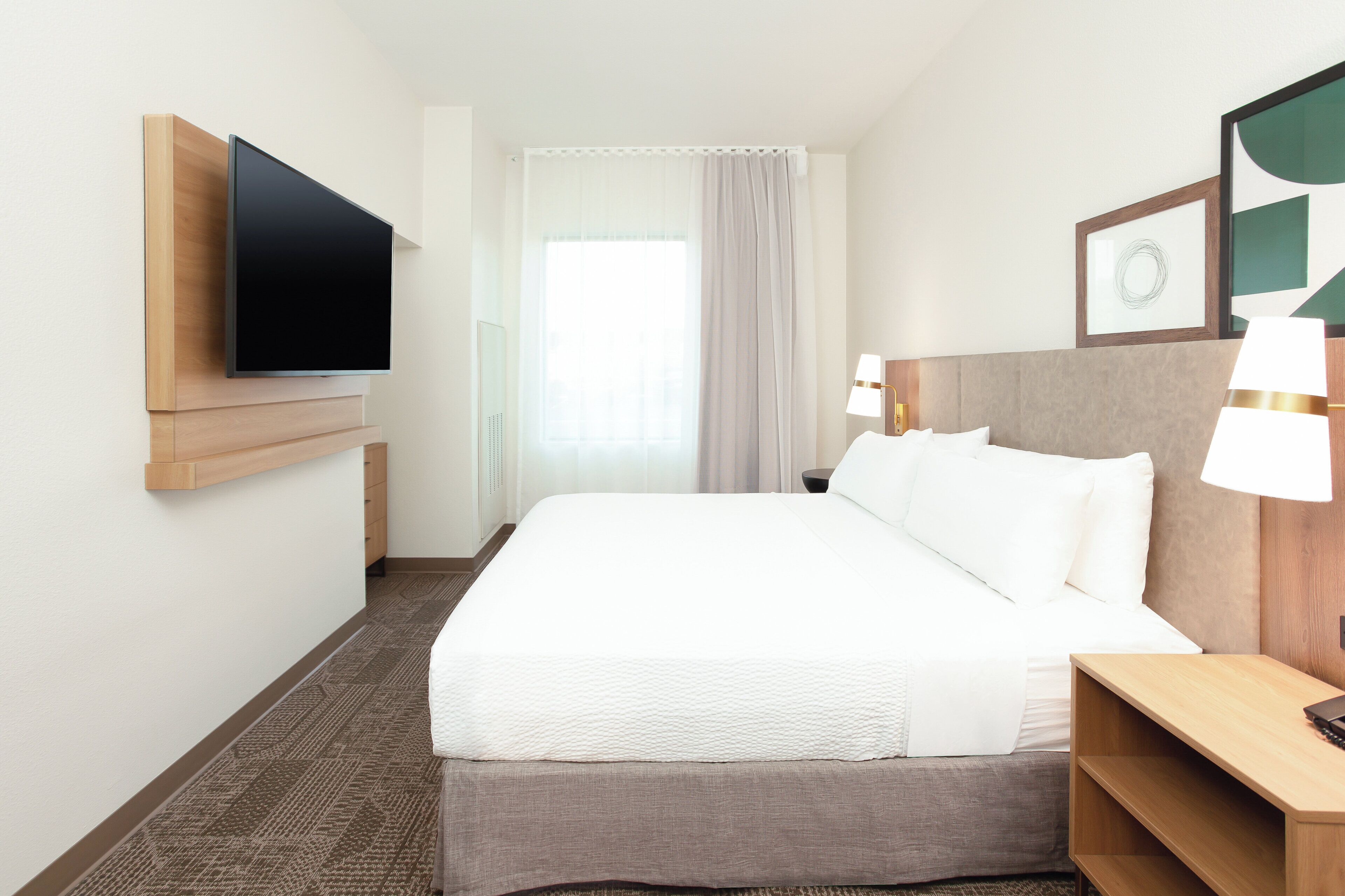 A hotel room with a large bed, a flat-screen TV mounted on the wall, a bedside table with a lamp, and a window with curtains.