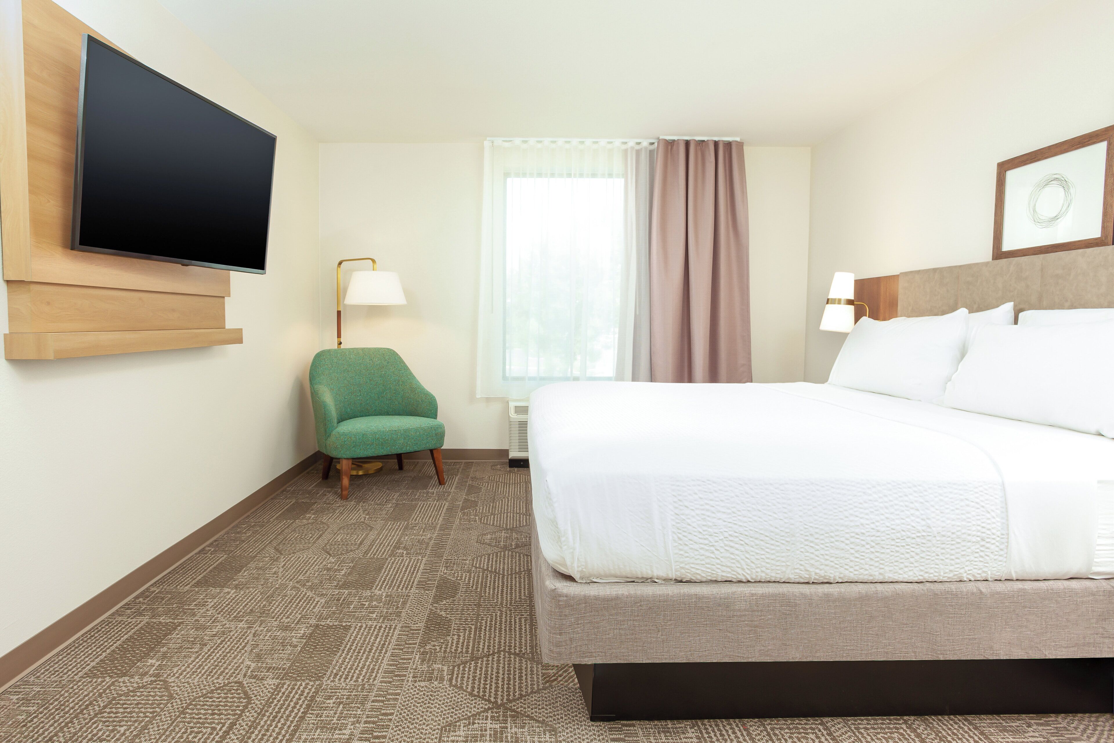 Quarto de hotel com cama, televisão na parede, cadeira, luminária e janela com cortinas.