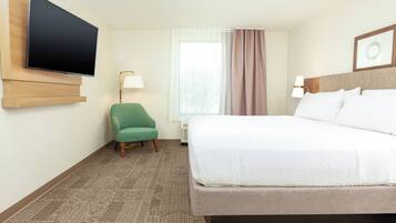 Quarto de hotel com cama, televisão na parede, cadeira, luminária e janela com cortinas.