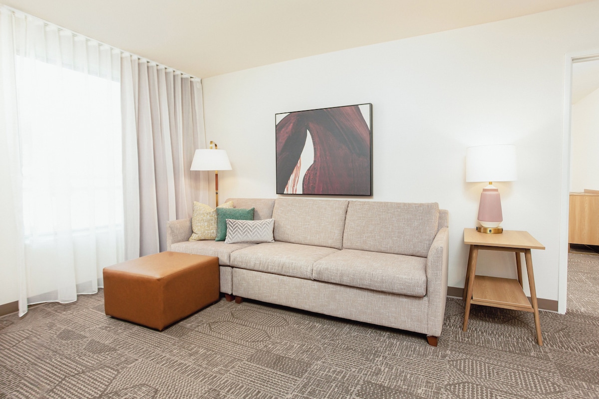 Una habitación de hotel con un sofá color beige, un reposapiés marrón, una mesita de madera con una lámpara y un cuadro enmarcado en la pared.