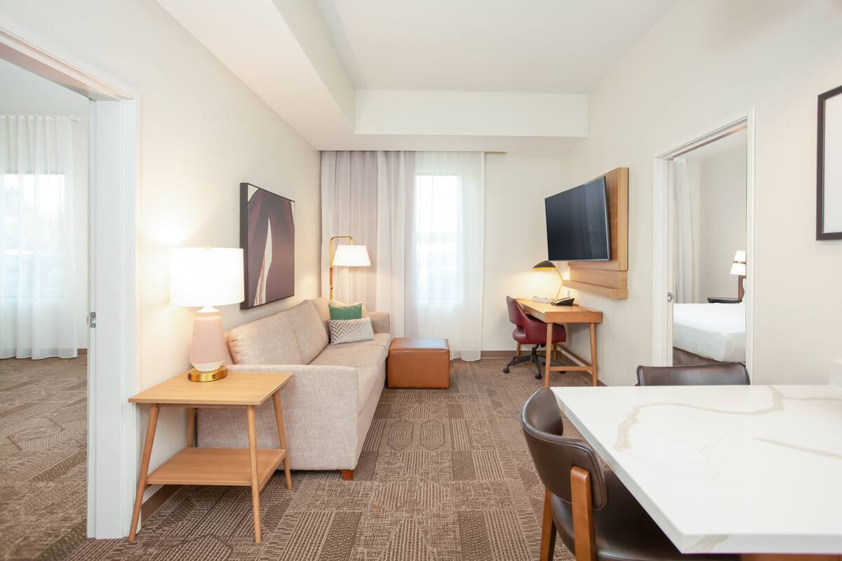 A hotel room with a sofa, a small table with a lamp, a desk with a chair, a TV mounted on the wall, and a bed in the background.