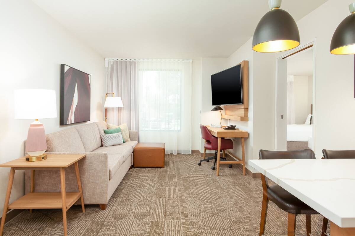 A modern hotel room with a sofa, a small table with a lamp, a desk with a chair, a TV, and a dining table with chairs.