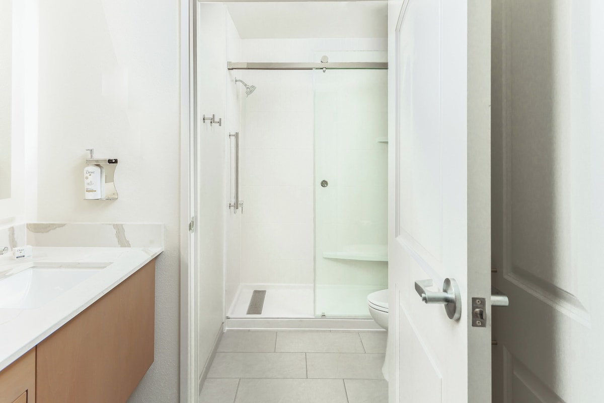 Un baño moderno con cabina de ducha de vidrio, un lavabo blanco y un tocador color beige.