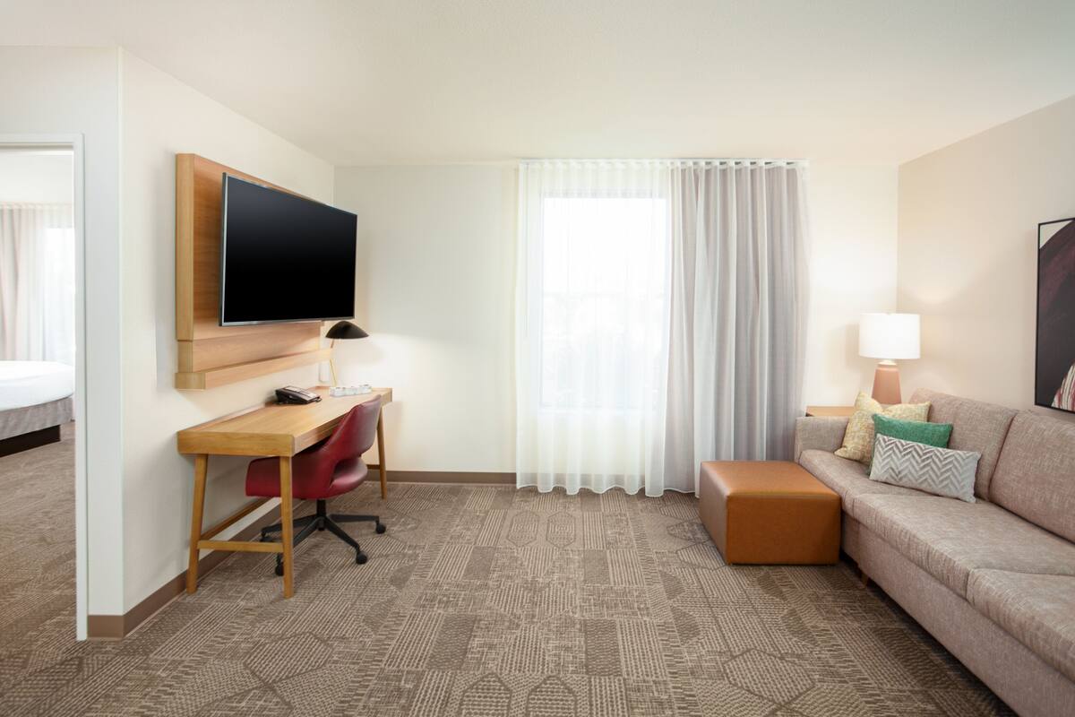 A hotel room with a wooden desk, a red office chair, a beige sofa, a brown ottoman, a flat-screen TV mounted on the wall, a window with white curtains, and a lamp on the sofa.
