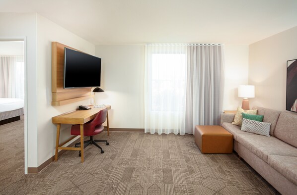 A hotel room with a wooden desk, a red office chair, a beige sofa, a brown ottoman, a flat-screen TV mounted on the wall, a window with white curtains, and a lamp on the sofa.