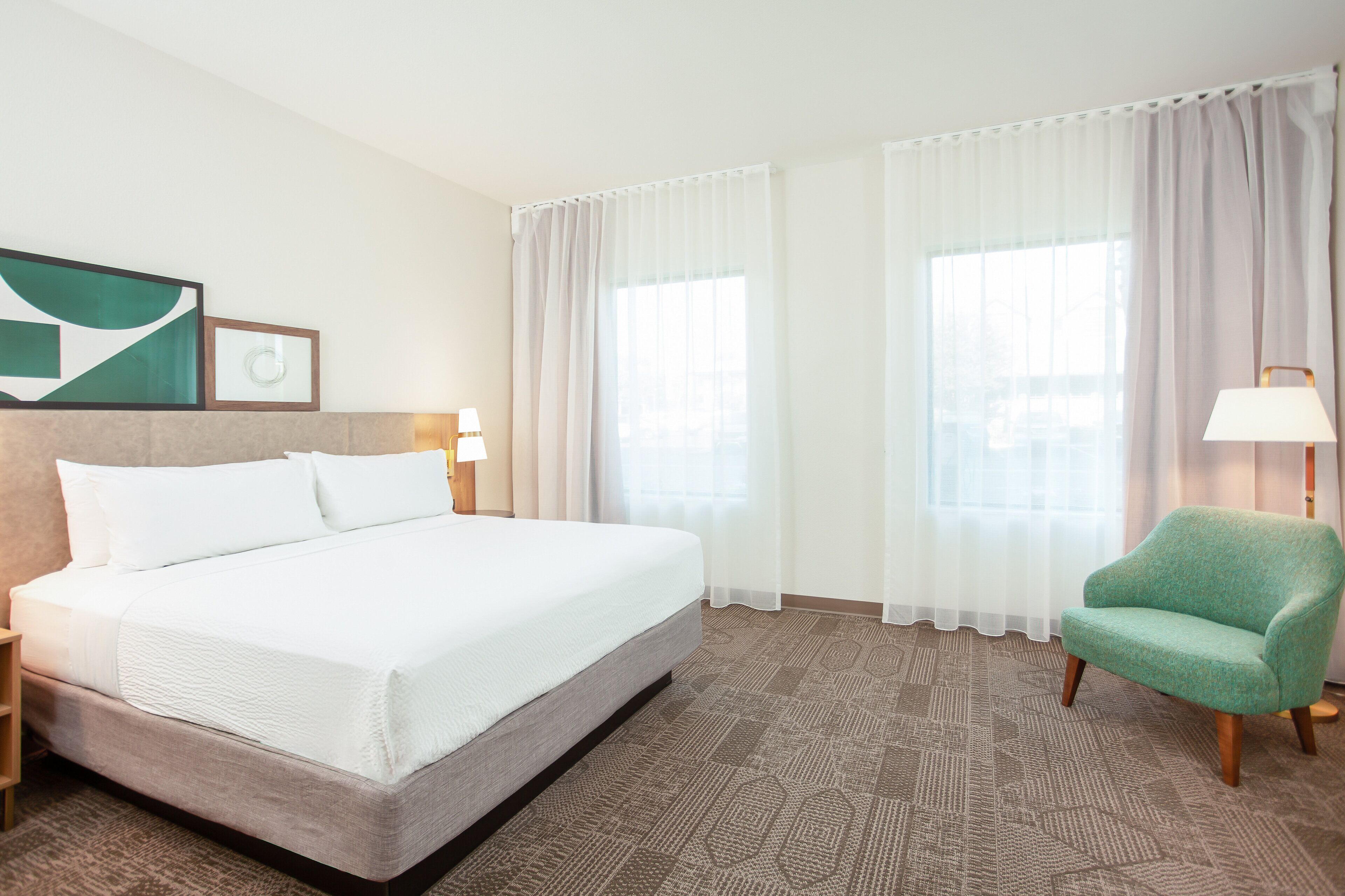 A hotel room with a large bed, two bedside tables with lamps, a green armchair, and a large window with sheer curtains.