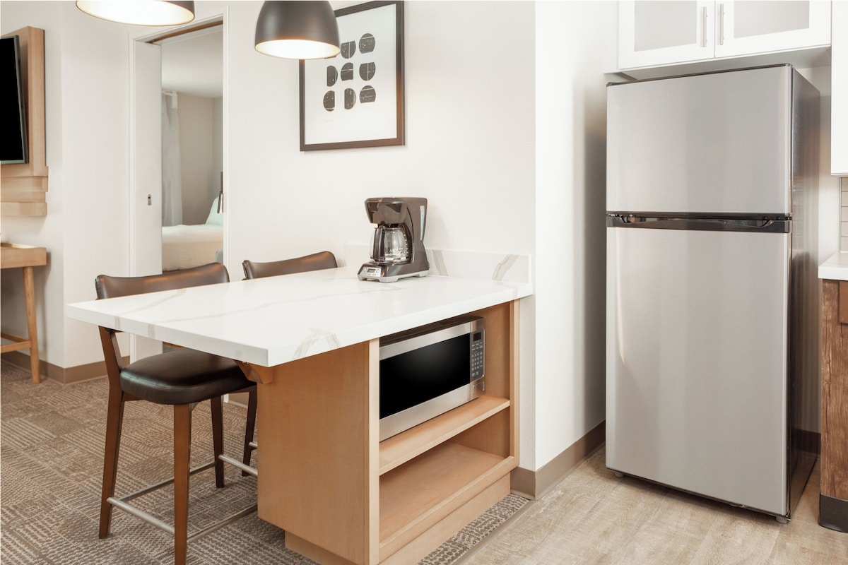 Cocina compacta con cubierta blanca, refrigerador de acero inoxidable, cafetera y microondas.