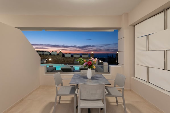 Un balcon donnant sur une piscine, avec une vue au coucher du soleil, une table avec des chaises et un vase rempli de fleurs.