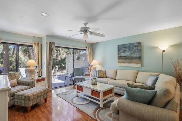 A spacious living room with a ceiling fan, a sofa set, a coffee table, and a view through a sliding glass door to an outdoor area.
