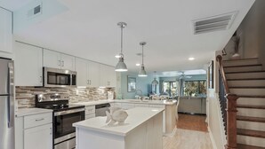 Cocina moderna con electrodomésticos de acero inoxidable, una isla central, iluminación colgante y vistas al salón.
