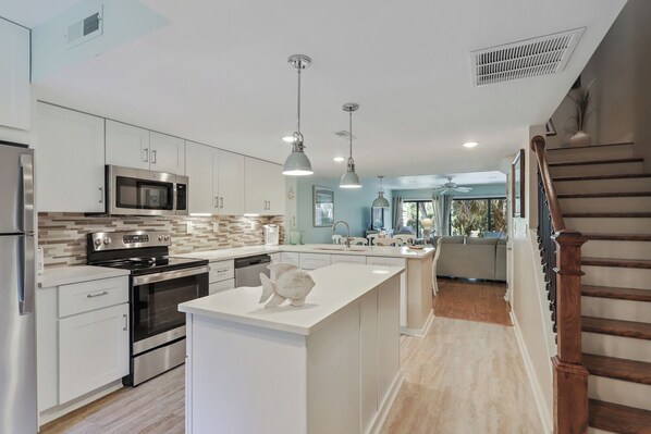 A modern kitchen with stainless steel appliances, a central island, pendant lighting, and a view to the living area.
