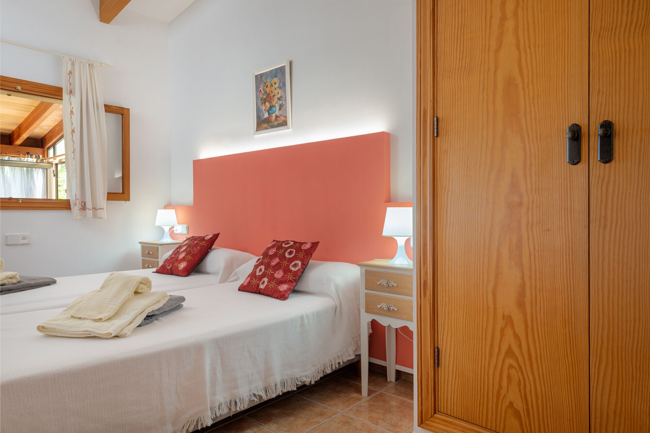 A bedroom with a bed, two bedside tables, a nightstand, a wooden wardrobe, a window with curtains, and a painting on the wall.