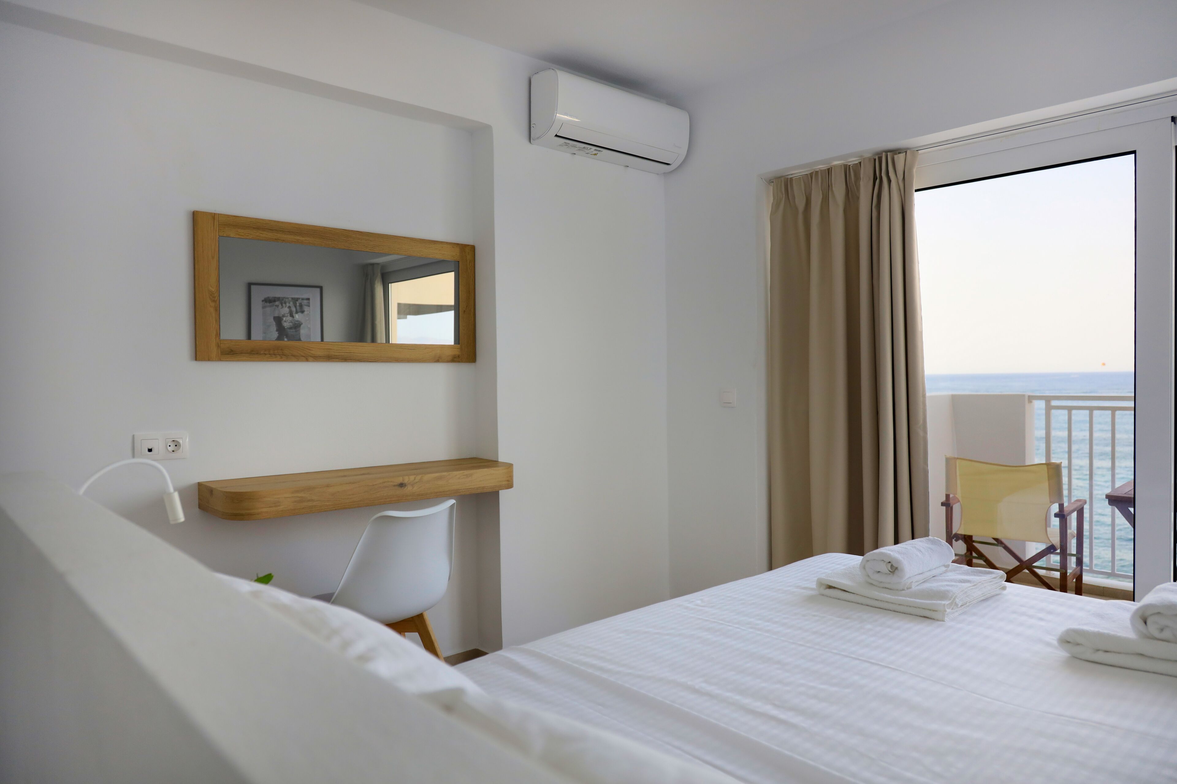 A hotel room with a bed, a wooden shelf, a desk, a chair, a mirror, and a view of the sea.