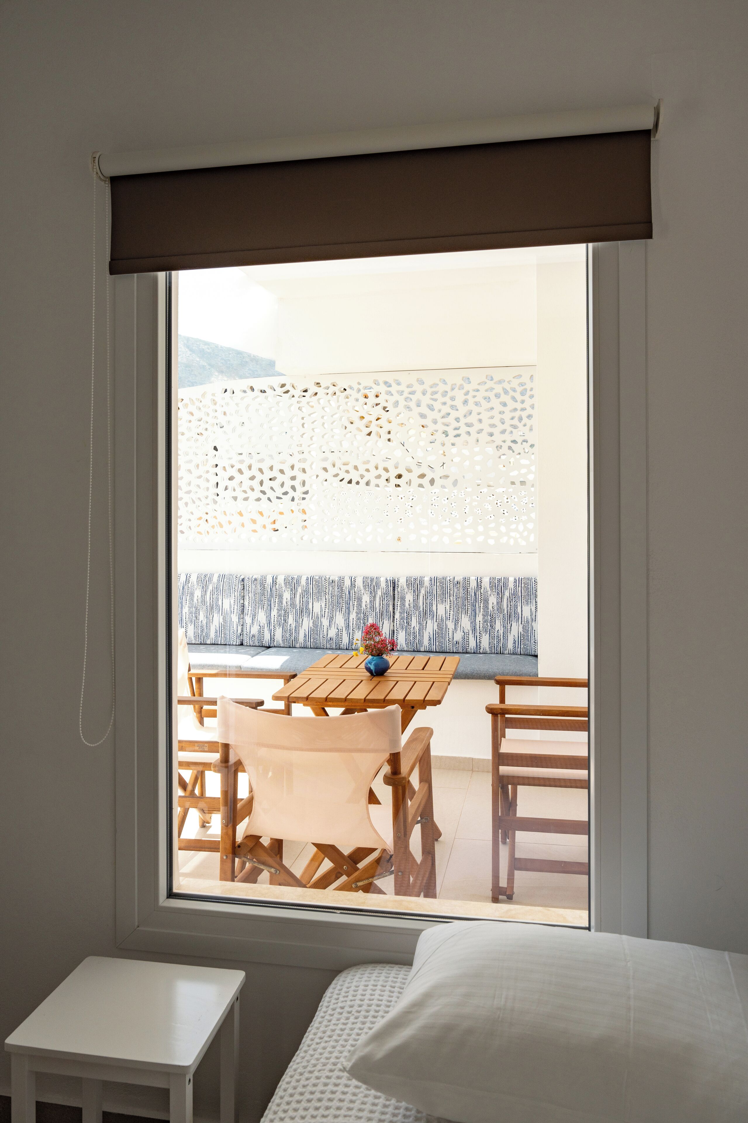 A room with a window, a small white table, a bed, and a view of an outdoor seating area with a patterned wall and a bench.