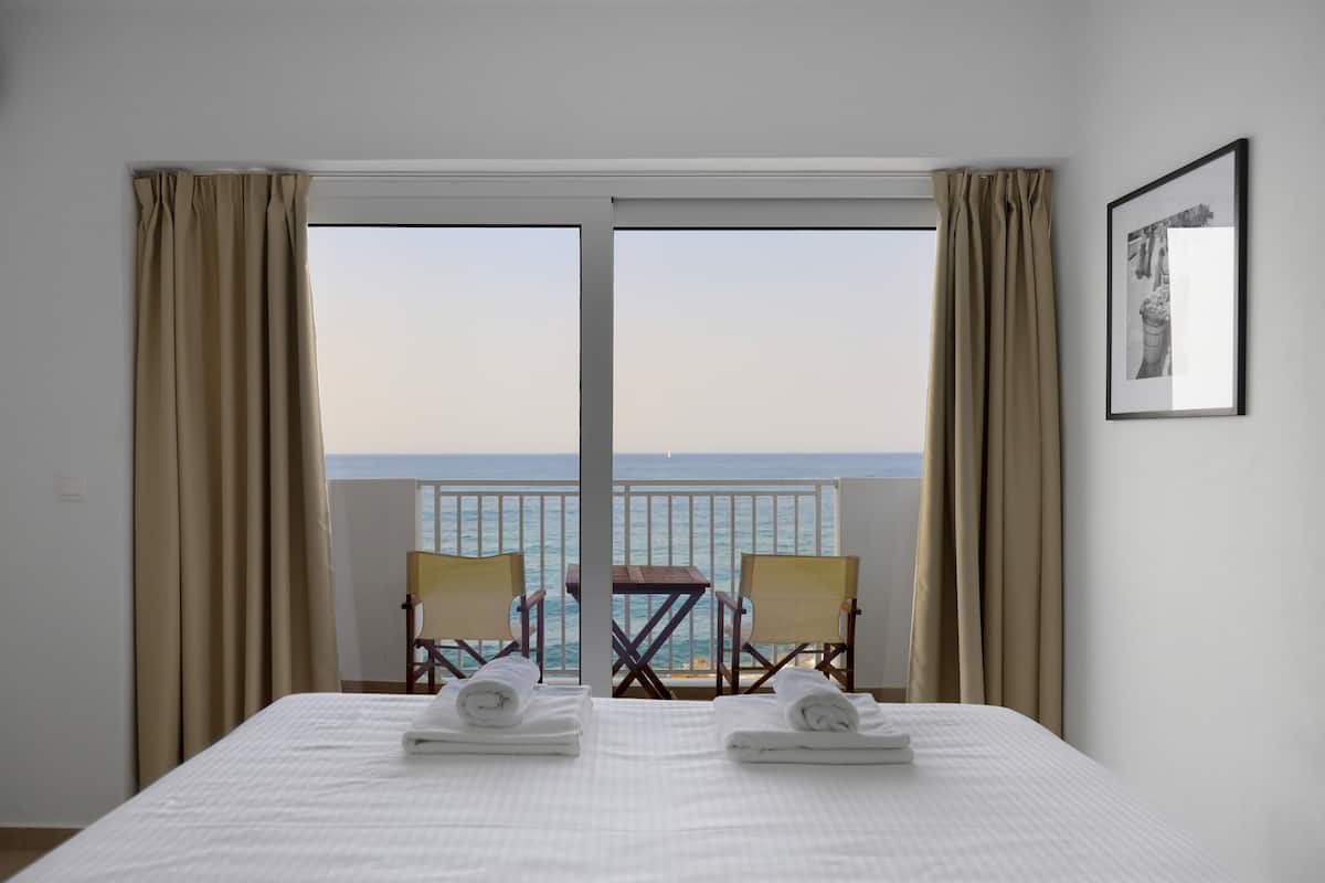 A hotel room with a large bed, two chairs, a small table, and a balcony with a view of the sea.