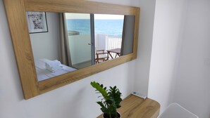 A hotel room with a wooden-framed mirror reflecting a balcony with a view of the sea. A potted plant is on a wooden table beside the bed. - Sea Breeze Hotel (Hersonissos)