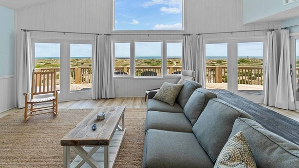 Een ruime kamer met een grote hoekbank, een houten tafel en uitzicht op het strand door grote ramen.
