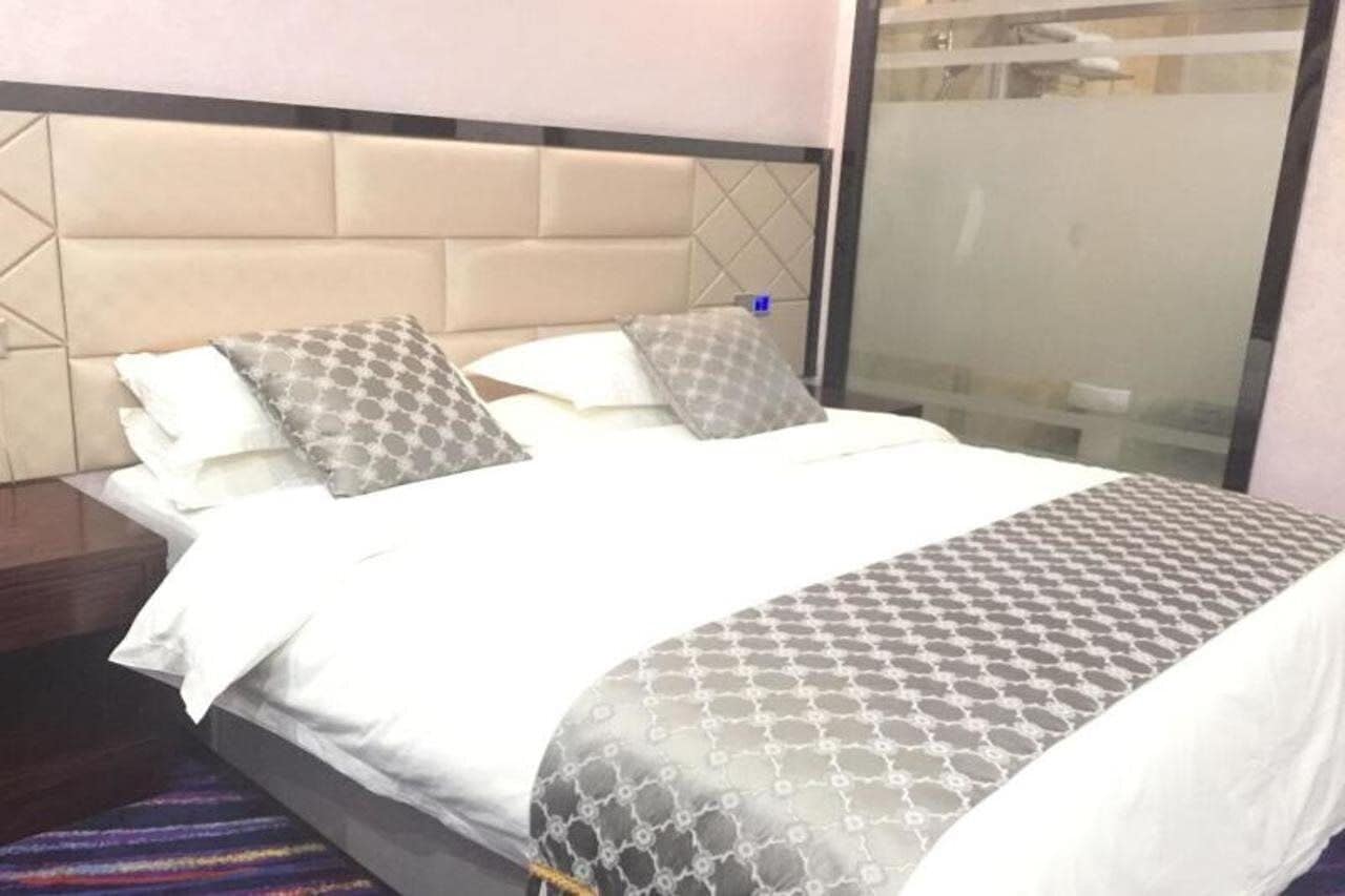 A neatly made bed with a patterned bedspread and pillows, a padded headboard, and a view of a bathroom through a glass partition.