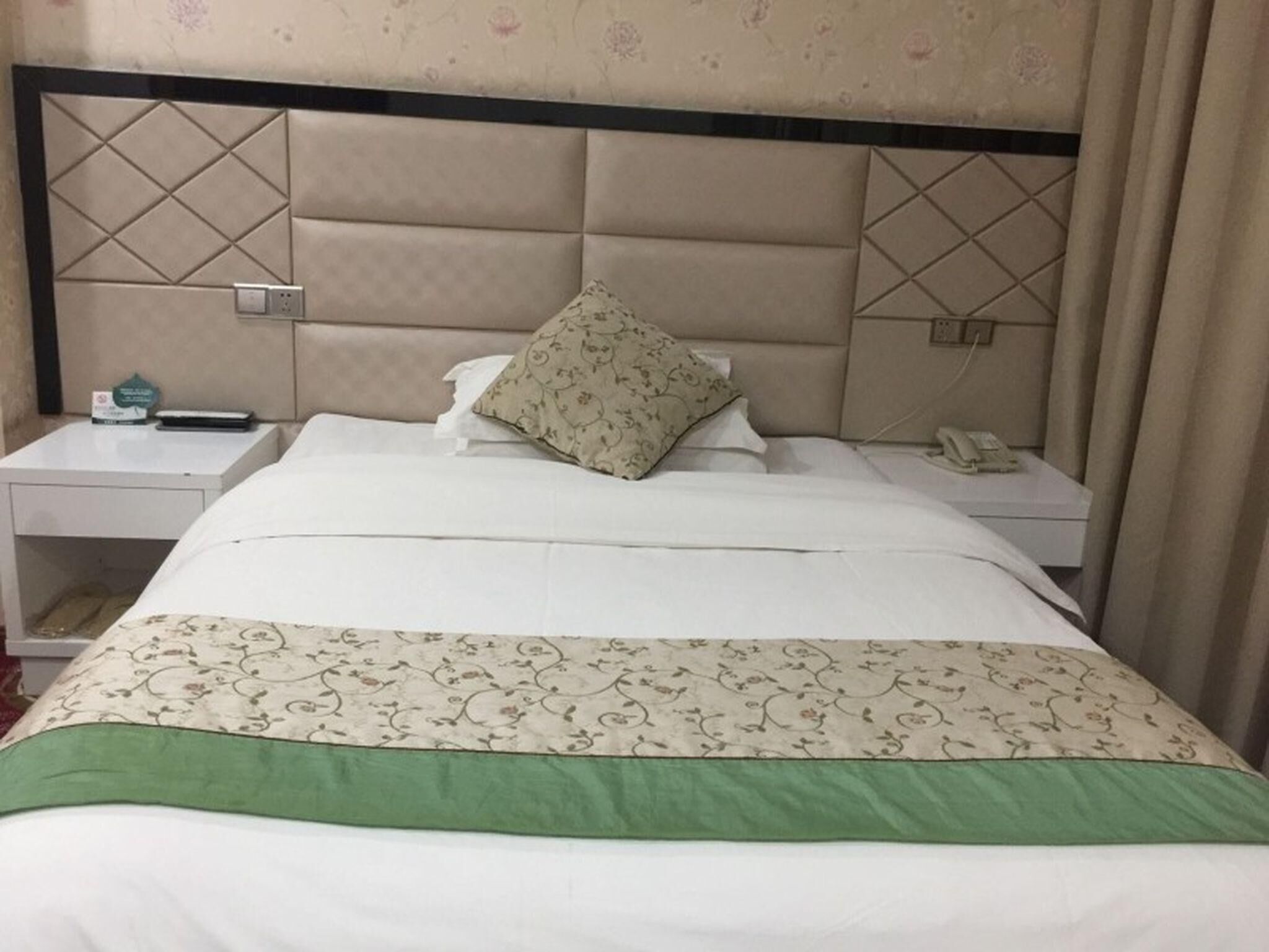 A hotel room with a padded headboard, a bed with a floral-patterned bedspread, a green accent at the foot of the bed, a nightstand with a phone and a small item on it, and a wall with a floral wallpaper pattern.