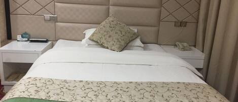 A hotel room with a padded headboard, a bed with a floral-patterned bedspread, a green accent at the foot of the bed, a nightstand with a phone and a small item on it, and a wall with a floral wallpaper pattern.