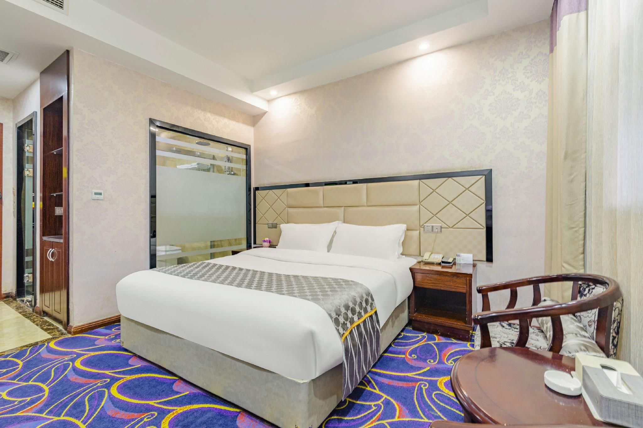 Una habitación de hotel con cama, mesita de noche, silla y baño con espejo y ducha.