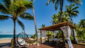 A wooden deck with a hanging chair and a canopy, overlooking a beach with palm trees and the ocean. - Vivant Eco Beach OFICIAL (Maraú)