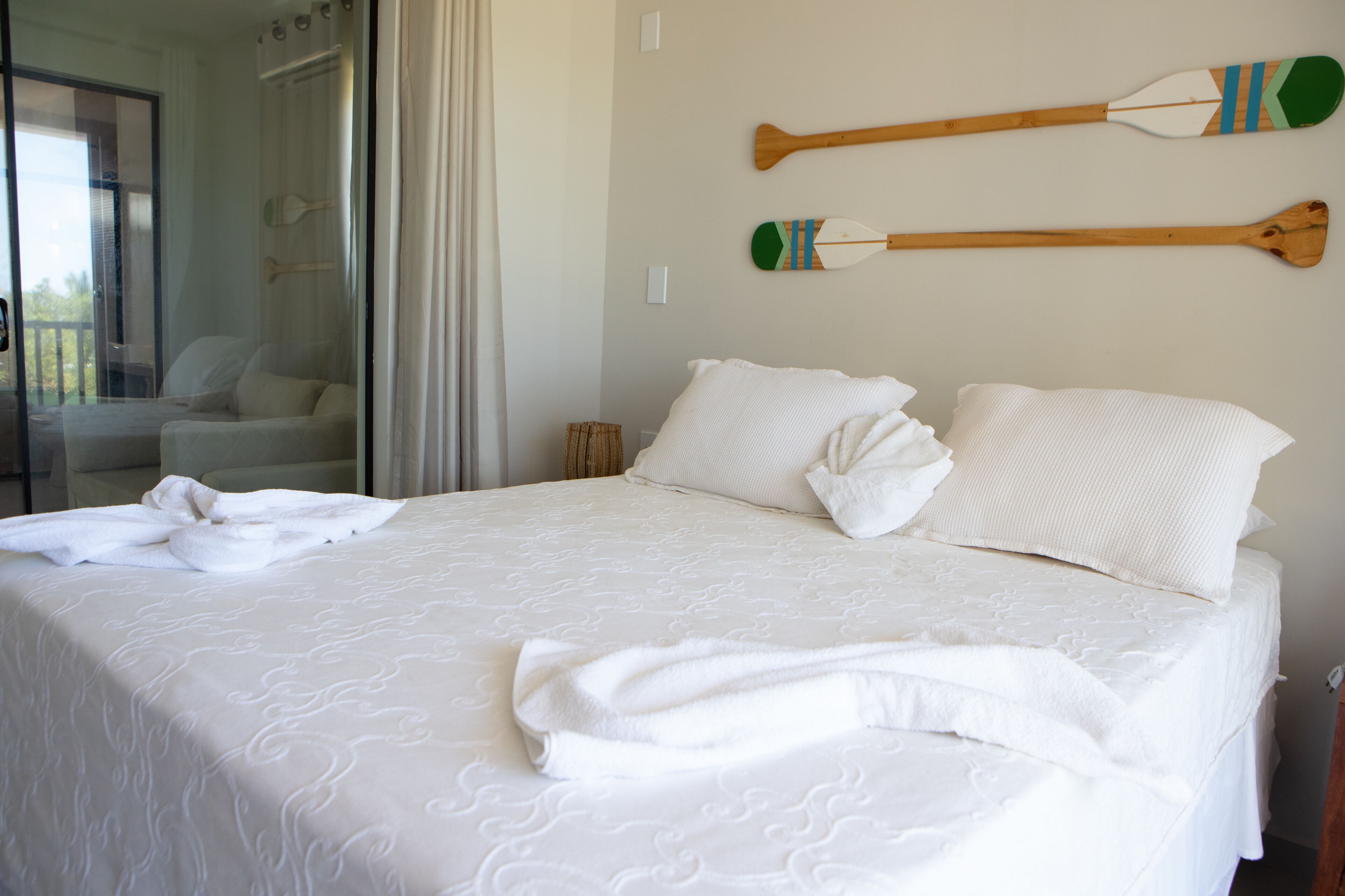 Cama bem arrumada com lençóis e almofadas brancas, decoração de parede com remo de madeira e vista para uma varanda com cadeira.