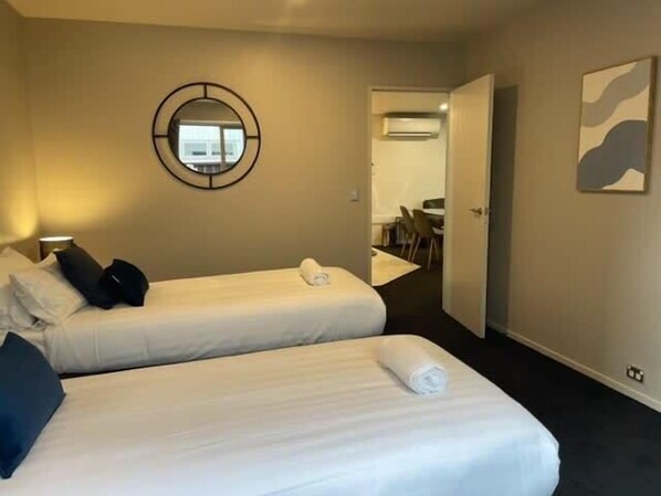 A hotel room with two beds, a round mirror, a painting, and an air conditioning unit in the background.