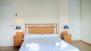 Una cama bien hecha con sábanas y toallas blancas, dos mesitas de noche con lámparas y un cabecero de madera.