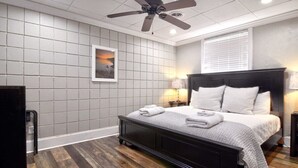 Un dormitorio con una cama de madera oscura, un ventilador de techo, un muro con azulejos grises, un cuadro enmarcado y una ventana con persianas.