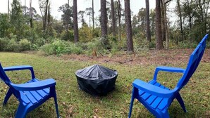 Dos sillas azules y un bote de basura negro en un área verde al aire libre con árboles de fondo.