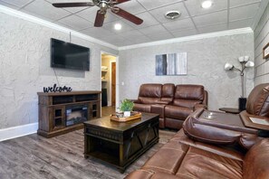 A living room with a leather sofa, a wooden coffee table, a TV mounted on the wall, and a ceiling fan.