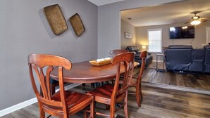 A dining area with a wooden table and chairs, a television mounted on the wall, and a ceiling fan. - The Cottage House - Family Friendly, Close to GSU! (Statesboro)