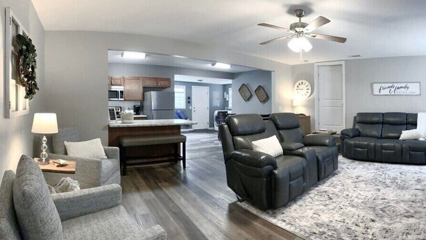 A modern living room with a gray sofa, a black leather recliner, and a gray armchair. A flat-screen TV is mounted on the wall, and a ceiling fan is visible. - The Cottage House - Family Friendly, Close to GSU! (Statesboro)