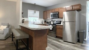 A modern kitchen with a central island, stainless steel appliances, wooden cabinets, and a grey floor. - The Cottage House - Family Friendly, Close to GSU! (Statesboro)