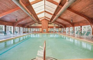 An indoor swimming pool with a high ceiling and wooden beams, surrounded by lounge chairs and large windows.