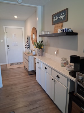 Una cocina con armarios blancos, encimera de madera, cafetera y una estantería con tazas.