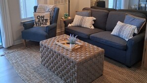 Un salon comprenant un canapé, un fauteuil et un pouf, une lampe et une fenêtre avec des stores.