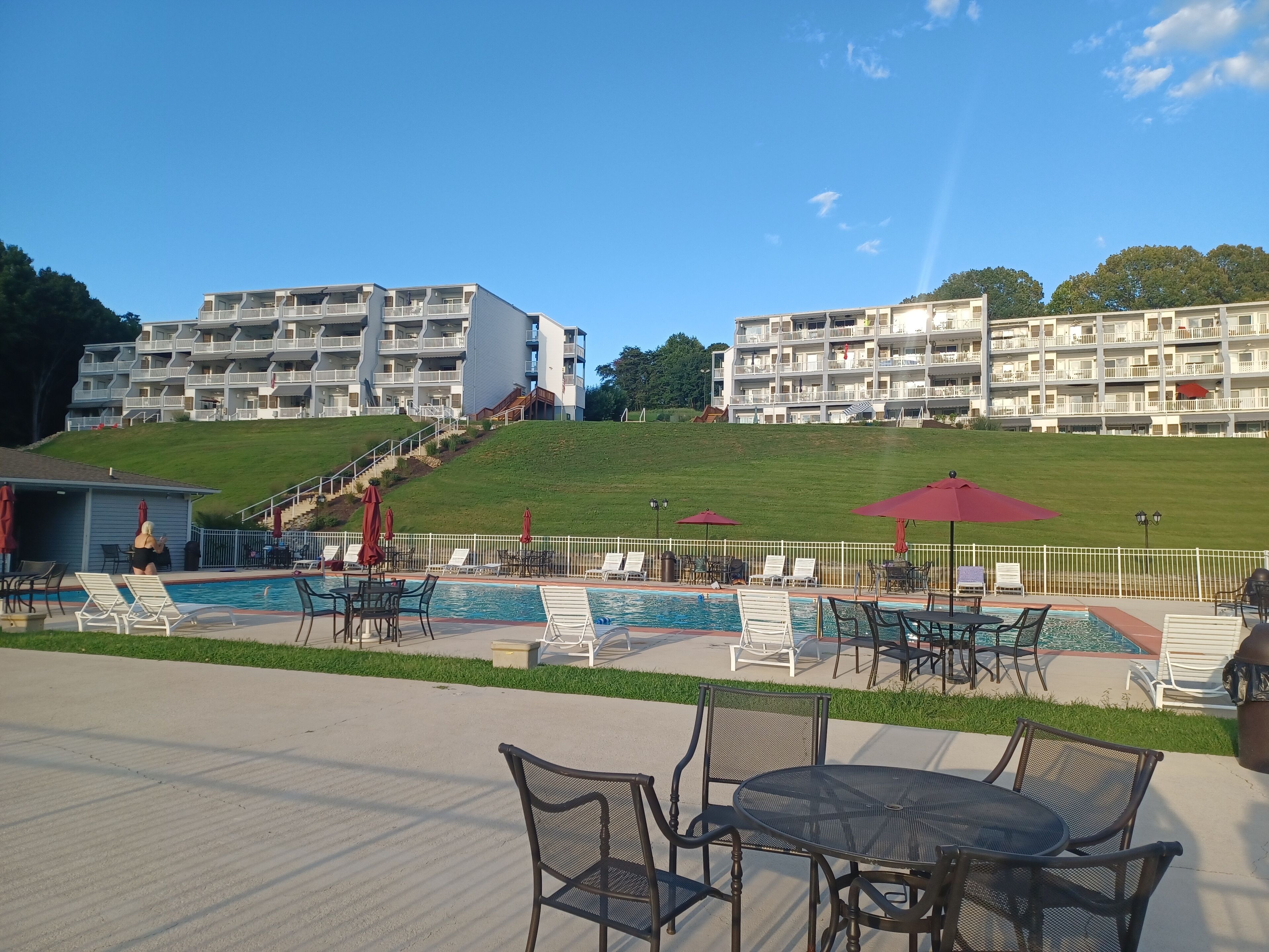 A pool area with lounge chairs and umbrellas, a building with multiple balconies, and a grassy hill in the background.