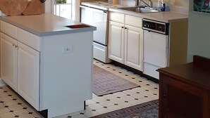 Une cuisine avec des meubles blancs, un îlot central, un tapis à motifs et un coin repas comprenant une table et des chaises en bois.