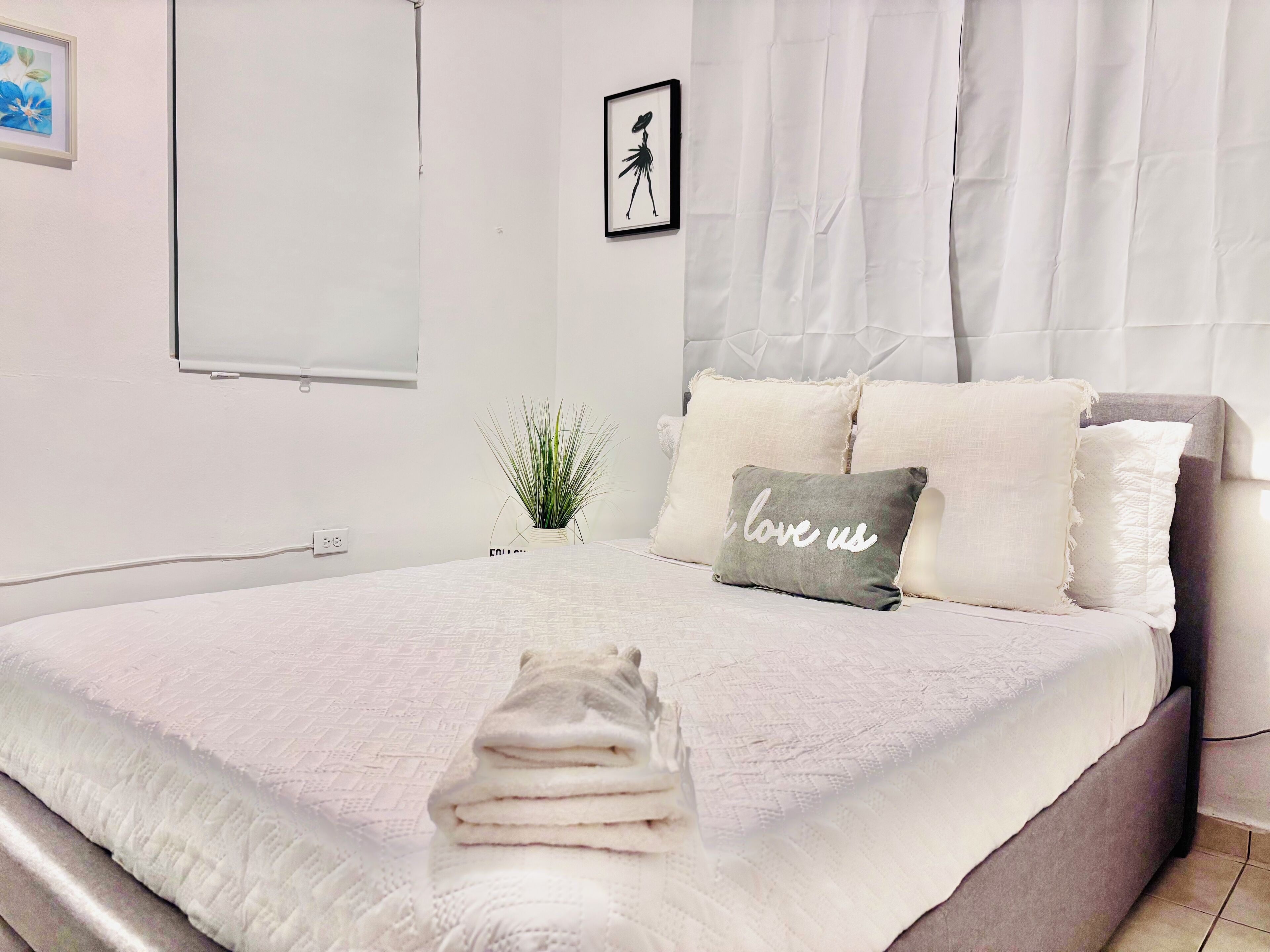 A neatly made bed with a folded towel, a small potted plant, and a pillow with "I love us" text.