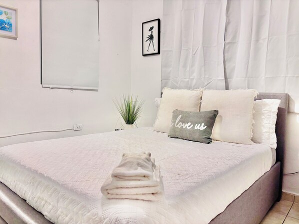 Una cama bien hecha con una toalla doblada, una pequeña planta en maceta y una almohada con el texto 'I love us'.