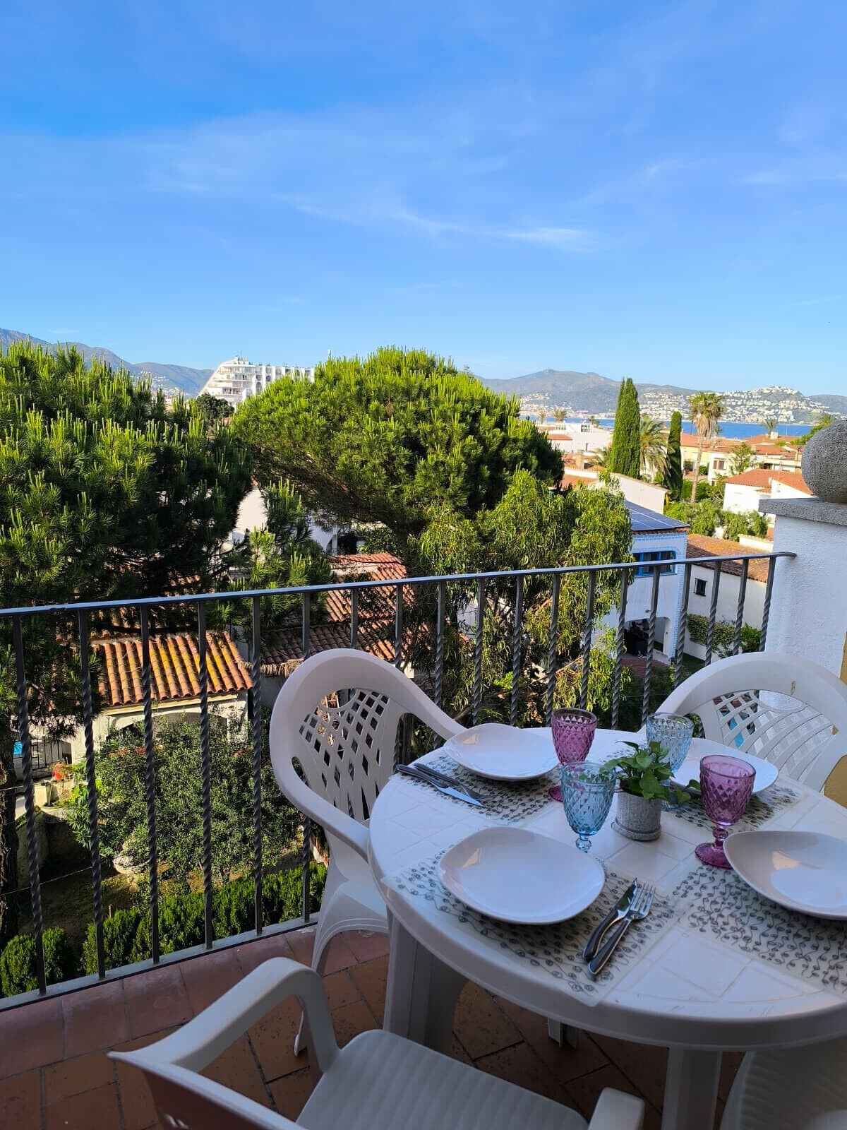 A balcony with a table set for two, overlooking a coastal town with buildings and trees.