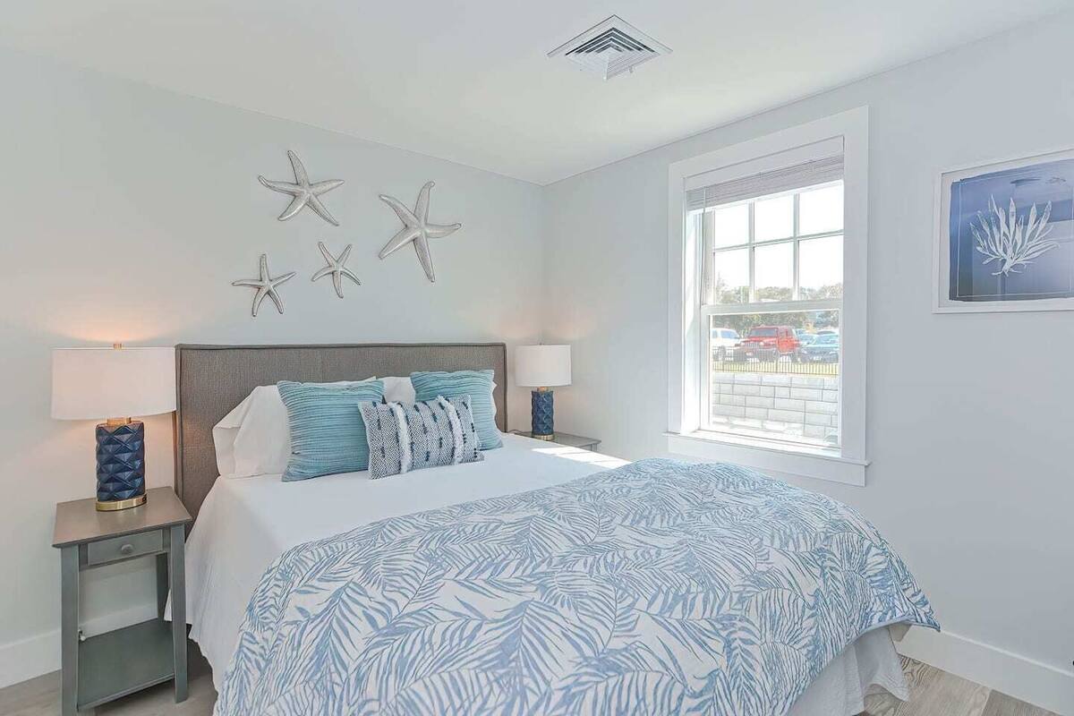 A bedroom with a bed, two bedside tables, starfish wall decor, a window with a view, and a ceiling vent.