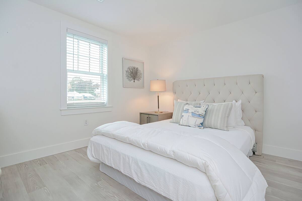 A bedroom with a large bed, a bedside table, a lamp, and a window with blinds.