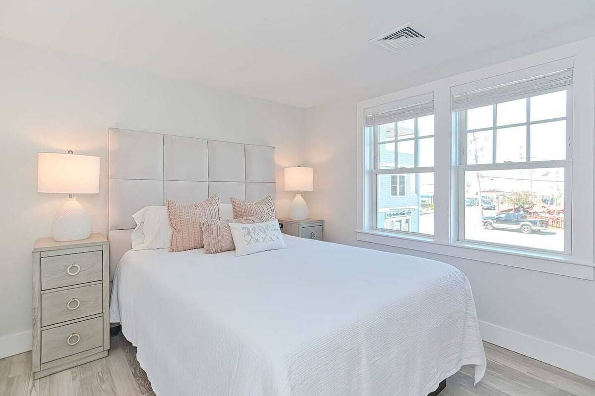 A bedroom with a large bed, two bedside tables with lamps, a window with a view of the street, and a ceiling vent.