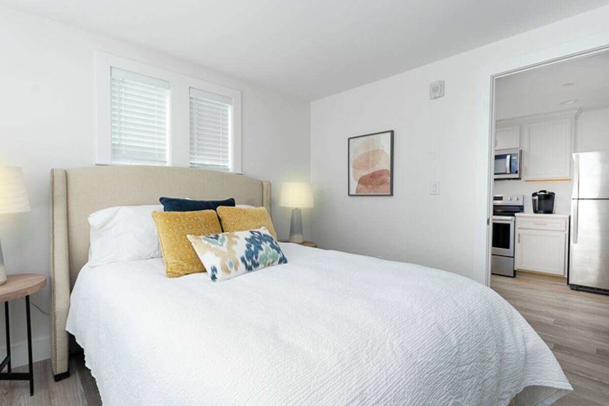 A bedroom with a bed, bedside lamps, a painting, and a view of a kitchen.
