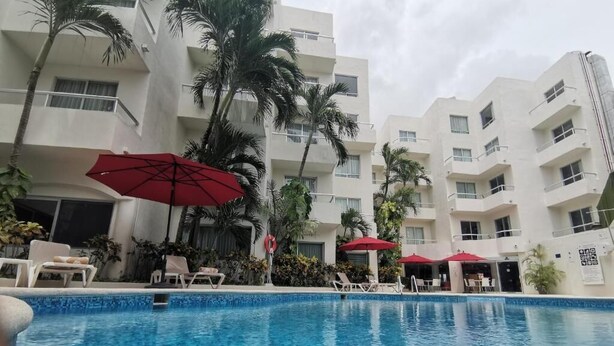 A swimming pool with lounge chairs and red umbrellas, surrounded by palm trees and a multi-story building. - Adhara Express (Cancun)