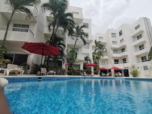A swimming pool with lounge chairs and red umbrellas, surrounded by palm trees and a multi-story building. - Adhara Express (Cancun)